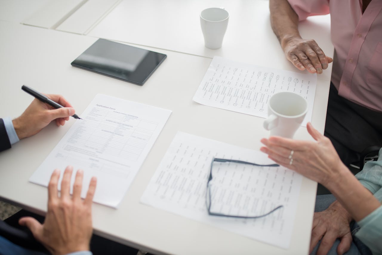 Hands writing on documents in a business meeting setup with coffee cups and tablet.